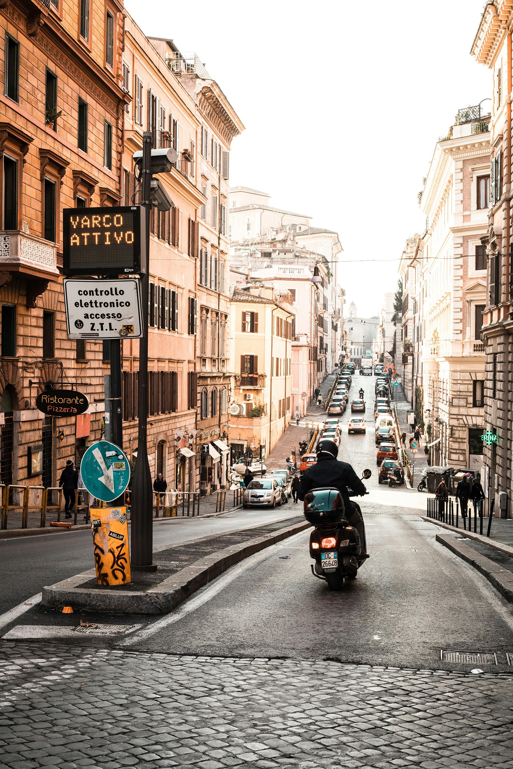 A person on a scooter rides up a busy, sunlit Rome street. The scene is framed by historic buildings, traffic, and a "Varco Attivo" sign. Urban, lively atmosphere.