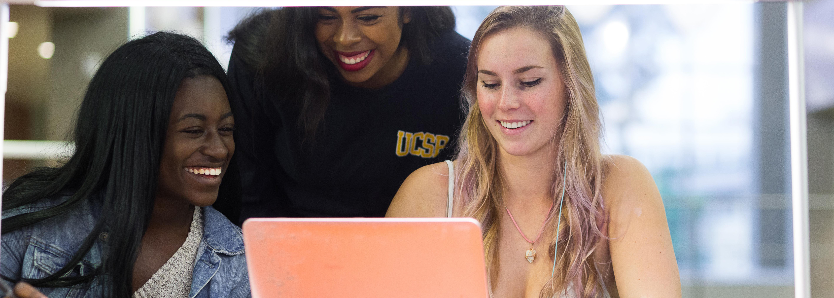 Three women smiling and looking at a laptop screen indoors. The scene conveys a joyful and collaborative atmosphere.